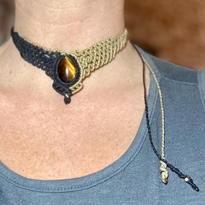 Może przedstawiać: Ręcznie robiony naszyjnik choker z makramy z owalnym kamieniem tygrysiego oka. Choker jest dwukolorowy, z czarnymi i beżowymi sekcjami makramy. Pasujący sznurek z makramy zwisa z naszyjnika.