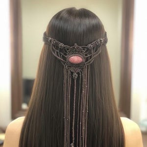 Può includere: Una fascia per capelli in macramè marrone con una gemma rosa ovale centrale e fili pendenti. L'intricato design presenta nodi decorativi e piccoli abbellimenti, creando un accessorio in stile bohémien o festival.