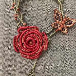 Può includere: Una collana in macramè caratterizzata da una grande rosa rossa arrotolata come elemento centrale con perline arancioni integrate, un piccolo motivo a farfalla con accenti viola e arancioni e un cordino verde oliva con dettagli a spina.