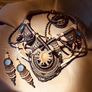Può includere: Un set di gioielli macramè fatti a mano con collana e orecchini abbinati. La collana ha un pendente centrale con una pietra arancione e dettagli blu. Gli orecchini hanno pietre blu e un design a strati. Il set è realizzato con cordoncini neri, beige e marroni.