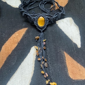 Può includere: Una collana girocollo in macramè nero con una pietra centrale a occhio di tigre ovale. La collana presenta dettagli a forma di foglie e un nappa di piccole pietre marroni e gialle, creando un'estetica bohémien.