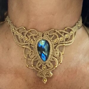 May include: A beige macrame choker necklace featuring an intricate knotted cord design with small gold-toned beads and a central teardrop-shaped labradorite gemstone that displays iridescent blue flashes against a dark stone background.