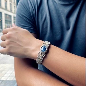 May include: A macramé bracelet featuring a central oval blue stone and detailed knotwork. The bracelet is made with a beige cord and gold-coloured accents. The design is visible on the wrist of a person wearing a blue t-shirt.