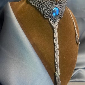 Può includere: Una collana macramè fatta a mano con una gemma blu ovale centrale. La collana presenta un'intricata annodatura in tonalità grigie e argentate, con un pendente intrecciato e una nappa. È esposta su un busto marrone.