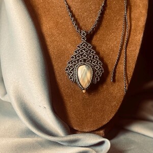 May include: A handcrafted macrame necklace with a teardrop-shaped pendant. The pendant features a light-colored stone with a unique pattern, set within a detailed, dark-colored macrame design. The necklace is displayed on a brown bust.