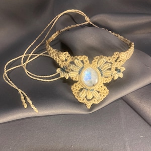 May include: A handmade macrame necklace featuring a central, iridescent stone. The necklace is crafted with beige cord, with intricate knotting details forming a floral design around the stone. The background is a dark gray fabric.