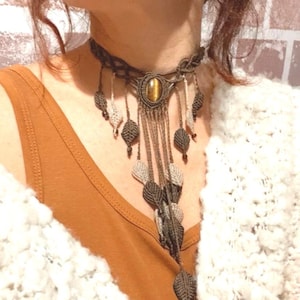 May include: A handcrafted macrame necklace with a central oval-shaped stone and leaf-shaped accents. The necklace features a brown and beige color scheme, with intricate knotting details. The necklace is a bohemian style accessory.