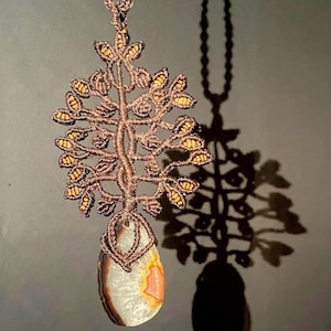 May include: A handcrafted macrame necklace featuring a tree design. The pendant has a brown macrame tree with yellow accents and a teardrop-shaped agate stone. The necklace is displayed against a dark background.