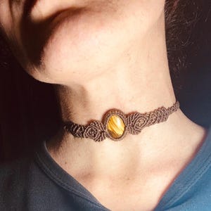 Peut inclure: Un collier ras du cou en macramé marron avec une pierre précieuse ovale jaune-brun au centre. Le collier, fait de cordon noué, est porté autour du cou. La pierre a un motif rayé. C'est un bijou fait à la main.