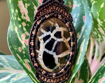 May include: A brown and black gemstone pendant with a white and brown pattern, set in a macrame knot with brown and copper cord and black, brown, and gold beads.