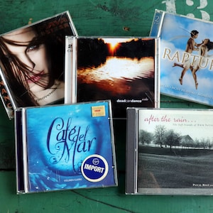 May include: Five compact discs with various album covers. The covers feature a woman with dark hair, a sunset over a lake, a couple dancing in the clouds, a blue ocean scene, and a rural landscape.