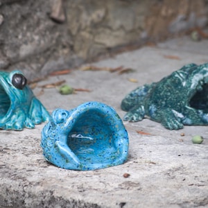 Può includere: Tre sculture di animali in ceramica che includono una rana color ottanio con occhi scuri, una rana blu brillante maculata e un alligatore verde scuro con la bocca aperta. Ogni pezzo è cavo, creando un design con un'ampia apertura.