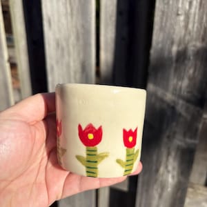 Könnte beinhalten: Ein kleiner, cremefarbener Keramikbecher mit handgemaltem Blumendesign. Das Design zeigt rote Tulpen mit gelben Zentren und grünen Stielen. Der Becher wird in einer Hand vor einem verwitterten Holzzaun gehalten.