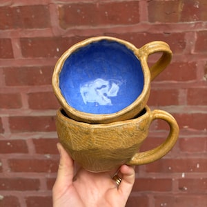 May include: Two handmade ceramic mugs stacked together. The exterior of the mugs is a textured, earthy brown, while the interior of the top mug is a vibrant blue. Each mug has a curved handle, perfect for holding hot beverages.