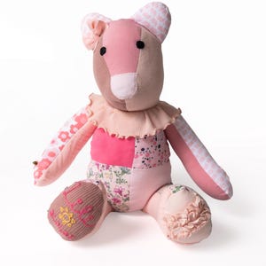 May include: A patchwork teddy bear constructed from various shades of pink and floral patterned fabric. The bear features a ruffled collar, a small bow on one ear, and embroidered text reading "Lilli" with a sun icon on its right foot.