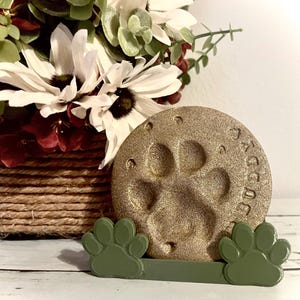 Puede incluir: Una piedra conmemorativa dorada con huella de pata y la palabra "Buddy" inscrita. La piedra está sostenida por una base verde en forma de pata. Un arreglo floral en una cesta tejida está en el fondo.