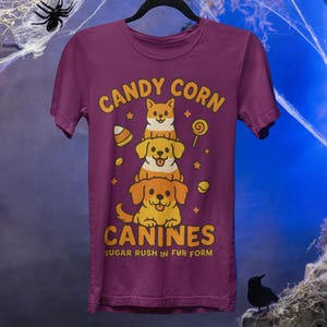 Può includere: Una maglietta viola con un'illustrazione di tre cani dei cartoni animati impilati, nei toni del giallo e dell'arancione. Il testo "CANDY CORN CANINES" e "SUGAR RUSH IN FUR FORM" è presente. Un design a tema Halloween.