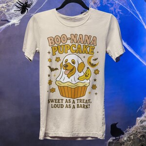 May include: Cream-colored t-shirt with a Halloween-themed graphic. The design features a dog dressed as a ghost inside a cupcake, with a banana, moon, and stars. Text reads "BOO-NANA PUPCAKE" and "SWEET AS A TREAT, LOUD AS A BARK!"