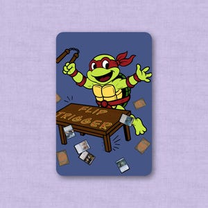 May include: A rectangular card with a blue background featuring a cartoon green turtle wearing a red mask and wristbands, holding nunchucks. The turtle stands behind a brown table with the text FLIP TRIGGER, surrounded by scattered playing cards.