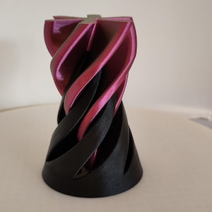 Könnte beinhalten: Ein 3D-gedrucktes dekoratives Objekt mit einem verdrehten, spiralförmigen Design. Der obere Teil ist in einem leuchtenden Magenta gehalten, während die Basis schwarz ist. Das Objekt hat eine glatte, glänzende Oberfläche und steht auf einer weißen Oberfläche.