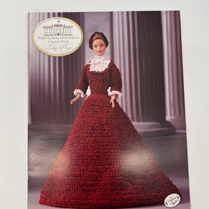 Può includere: Bambola della collezione First Ladies of America, con un abito all'uncinetto rosso scuro con finiture in pizzo bianco. La bambola ha i capelli castani e posa davanti a uno sfondo sfocato di colonne.