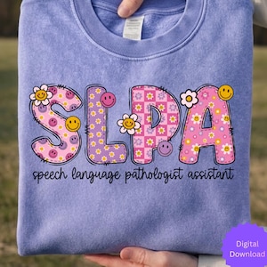 Könnte beinhalten: Ein helllila Sweatshirt mit den Buchstaben "SLPA" in einem farbenfrohen, floralen Design. Die Buchstaben sind mit Smiley-Gesichtern und Blumen verziert. Der Text "speech language pathologist assistant" ist darunter gedruckt.