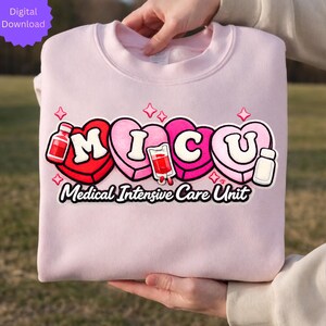 May include: A light pink sweatshirt with the text "MICU" in a heart-shaped design, with medical-themed illustrations. The words "Medical Intensive Care Unit" are written below the design in a curved font.