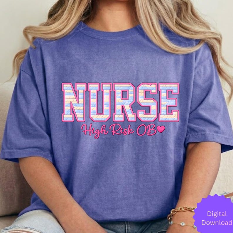 May include: A periwinkle blue t-shirt with the word "NURSE" in a pink and pastel-colored block letter design. Below the word "NURSE" is the text "High Risk OB" with a pink heart. The shirt is a casual style.