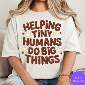 May include: Cream-colored t-shirt with the phrase "HELPING TINY HUMANS DO BIG THINGS" in brown, textured letters. The design includes small star and heart accents. The shirt is a casual style.