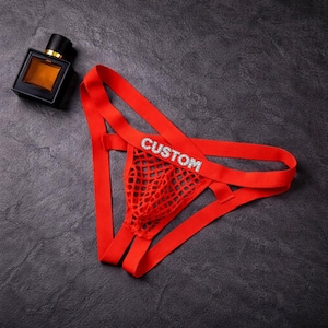 May include: Red thong underwear with a mesh front panel and the word "CUSTOM" spelled out in rhinestones. The underwear has a minimalist design with thin straps. A black perfume bottle with a gold cap is in the background.