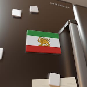 May include: A rectangular magnet featuring the Iranian flag, with green, white, and red stripes, and a golden lion and sun emblem. The magnet is attached to a dark refrigerator door, alongside other white magnets.