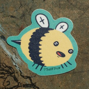 May include: A yellow and black bee sticker with a surprised expression. The bee has a turquoise outline and two X's on its head. The sticker says "Toady Town" at the bottom.