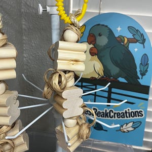 Puede incluir: Un juguete para pájaros hecho de bloques de madera natural, cuentas de madera y material tejido, suspendido de un anillo de plástico amarillo. El juguete cuelga de un expositor de metal blanco. Un letrero azul con ilustraciones de loros está en el fondo.