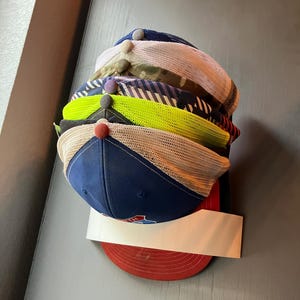 May include: A stack of baseball caps in various colors, including navy blue, red, and neon green. The caps have mesh backs and different colored brims. The caps are stacked on top of each other, with a white label visible.