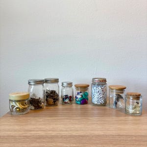 May include: Eight clear glass jars of varying sizes arranged in a row. The jars feature metal or wooden lids and contain assorted craft supplies including white rings, metal findings, colorful buttons, letter beads, and small metallic objects.