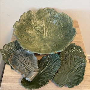 Op de afbeelding: Een grote, ronde, groene keramische kom in de vorm van een koolblad, vergezeld van vier kleinere, individuele groene keramische borden die ook de vorm hebben van koolbladeren, allemaal met gedetailleerde, reliëf nerven en een glanzende afwerking.