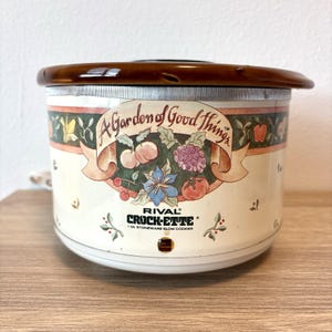 May include: A Rival Crock-Ette 1 litre stoneware slow cooker with a cream-coloured exterior featuring a decorative fruit and vegetable illustration, a banner with the text A Garden of Good Things, and a dark brown lid with a glossy finish.