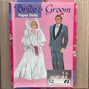 Könnte beinhalten: Ein rosafarbener rechteckiger Karton für Bride & Groom Papierpuppen. Die Vorderseite zeigt Illustrationen einer Braut im weißen Kleid und eines Bräutigams im blauen Anzug. Ein kleineres Bild zeigt die Puppen und Outfits. Der Text lautet Bride & Groom.