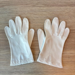 Peut inclure: Une paire de gants blancs arrivant au poignet, fabriqués dans un tissu lisse avec un bord festonné. Les poignets sont ornés de perles blanches délicates disposées selon un motif répétitif le long de l'ourlet inférieur de chaque gant.