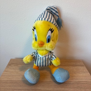 May include: A plush Tweety Bird character wearing a blue and white striped nightcap with a matching tassel and a blue and white striped pajama top. The figure has bright yellow fur, large blue eyes with long black eyelashes, and orange feet.