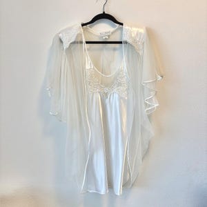 May include: A white satin chemise and matching sheer robe set. The chemise features a scoop neckline with white floral lace detailing. The robe has wide, flowing sleeves with lace accents at the shoulders and a delicate trim along the edges.