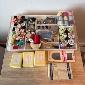 Könnte beinhalten: Ein weißer Kunststoff-Organizer gefüllt mit Nähzubehör wie bunten Garnrollen, Nadeln, Stecknadeln, Fingerhüten und einer Schere. Darunter befinden sich drei gelbe Schachteln mit T-Pins, eine Packung Talon-Nahtband und eine Needling-Schachtel.