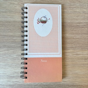 May include: A spiral-bound notebook featuring a peach-colored cover with a white rectangular panel containing a small oval illustration of a flower basket. The bottom section of the cover is a solid peach color with the word Notes printed in white.