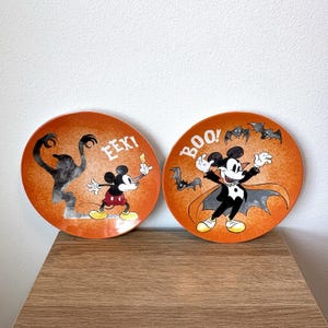 May include: Two round orange ceramic plates featuring Mickey Mouse. The left plate shows Mickey with a ghost and the text EEK!. The right plate depicts Mickey dressed as a vampire with bats and the text BOO!. Both plates have a speckled orange background.