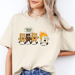 May include: Beige t-shirt with a cartoon graphic of s'mores. Three s'mores are chasing a burning marshmallow, with a speech bubble saying "GROUP HUG!!" The shirt is paired with blue jeans.