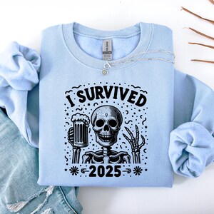 Puede incluir: Sudadera azul claro con un dise&ntilde;o en blanco y negro de un esqueleto sosteniendo una jarra de cerveza. El texto "I SURVIVED 2025" est&aacute; impreso encima y debajo del esqueleto. Un collar plateado est&aacute; colocado sobre la sudadera.
