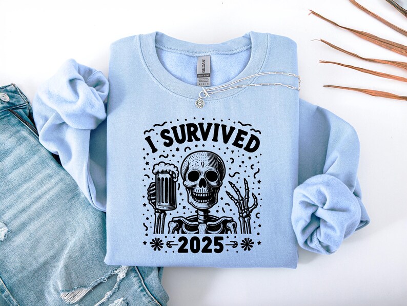 Puede incluir: Sudadera azul claro con un dise&ntilde;o en blanco y negro de un esqueleto sosteniendo una jarra de cerveza. El texto "I SURVIVED 2025" est&aacute; impreso encima y debajo del esqueleto. Un collar plateado est&aacute; colocado sobre la sudadera.