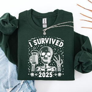 Puede incluir: Una sudadera verde oscuro con el texto "I Survived 2025" y un gr&aacute;fico de esqueleto sosteniendo una jarra de cerveza. Un collar plateado est&aacute; sobre la sudadera, que est&aacute; sobre unos vaqueros azules y una manga verde enrollada.
