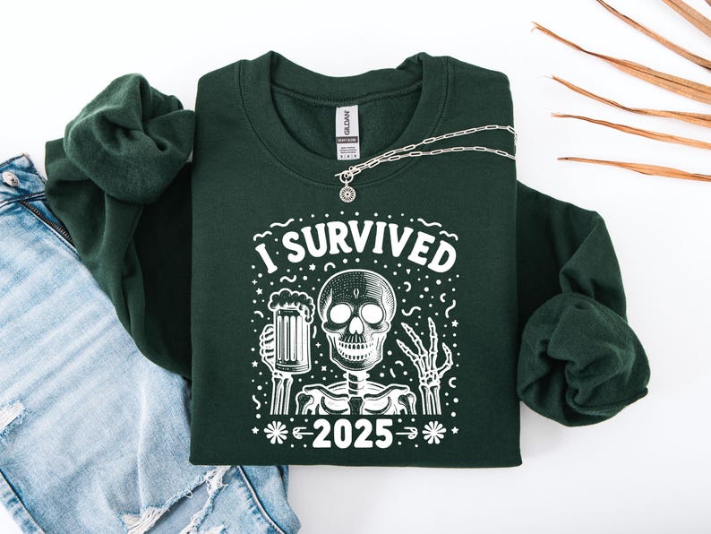 Puede incluir: Una sudadera verde oscuro con el texto "I Survived 2025" y un gr&aacute;fico de esqueleto sosteniendo una jarra de cerveza. Un collar plateado est&aacute; sobre la sudadera, que est&aacute; sobre unos vaqueros azules y una manga verde enrollada.