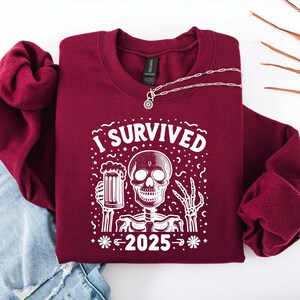Puede incluir: Sudadera burdeos con el texto blanco "I SURVIVED 2025". El gr&aacute;fico presenta un esqueleto sosteniendo una jarra de cerveza. Un collar plateado con un colgante est&aacute; sobre la sudadera. La sudadera est&aacute; sobre unos vaqueros.